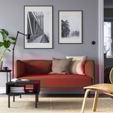 Modernes Wohnzimmer mit rotem LILLEHEM-Sofa, gemusterten Kissen, schwarzem Couchtisch, Kunstdrucken, Stehlampe und Topfpflanze.