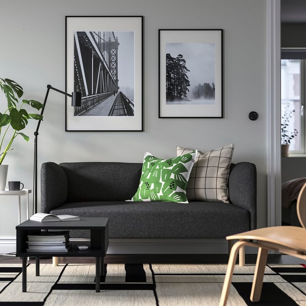 Modernes Wohnzimmer mit grauem Sofa, grünen/neutralen Kissen, schwarzen Fotos, Beistelltisch mit Büchern, Stuhl und Pflanze.