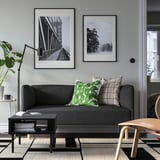 Modernes Wohnzimmer mit grauem Sofa, grünen/neutralen Kissen, schwarzen Fotos, Beistelltisch mit Büchern, Stuhl und Pflanze.