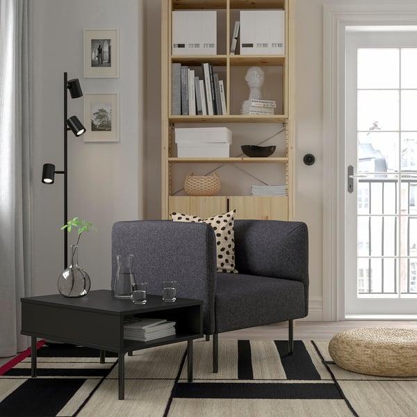 Modernes Wohnzimmer mit grauem LILLEHEM-Sofa, schwarzem Couchtisch, hölzernem Bücherregal, stilvollem Teppich und Tageslicht. Effizientes und gemütliches Design.