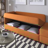 Orangefarbenes Schlafsofa mit offenem Staufach mit Kissen. Karierter Teppich, Beistelltisch mit Snacks.