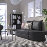 Modernes Wohnzimmer mit grauem LINDÅKRA-Schlafsofa, gemustertem Teppich, Bücherregal, Couchtisch und Dekor.
