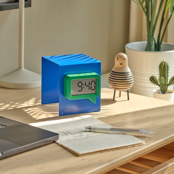 Eine blaue Würfeluhr mit grüner Sprechblasenanzeige um 9:40 Uhr auf einem Holztisch mit Notizbuch, Stift, Laptop und dekorativen Gegenständen in der Nähe.
