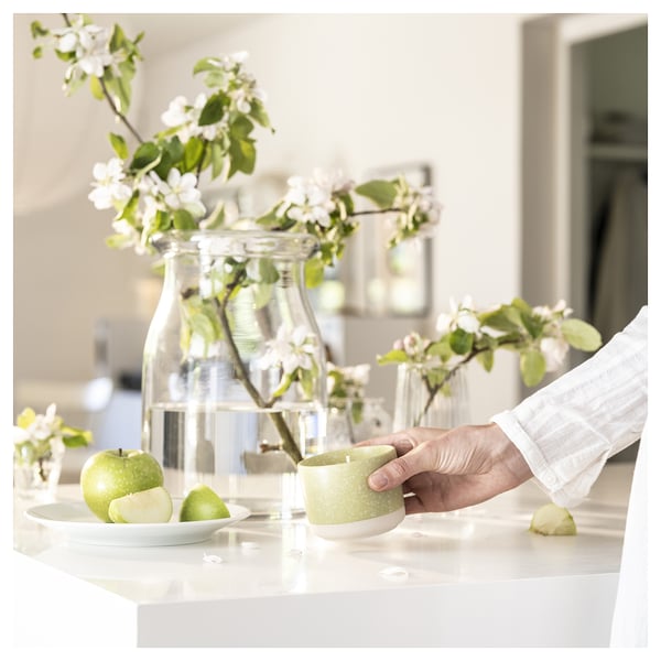 Eine Person hält eine grüne LÖVSKOGSLUND-Duftkerze neben Äpfeln und Blumen.
