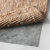 Ein Bild, das einen beigen Juteteppich mit rutschfestem Filzrücken zeigt. Der Teppich ist gefaltet, um seine Unterseite zu zeigen, wodurch das funktionale Filzmaterial hervorgehoben wird.