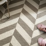 Gestreifter Wollteppich mit beigen und grauen Streifen. Rosa Sandalen obendrauf, Stuhlbein aus Metall zu sehen.