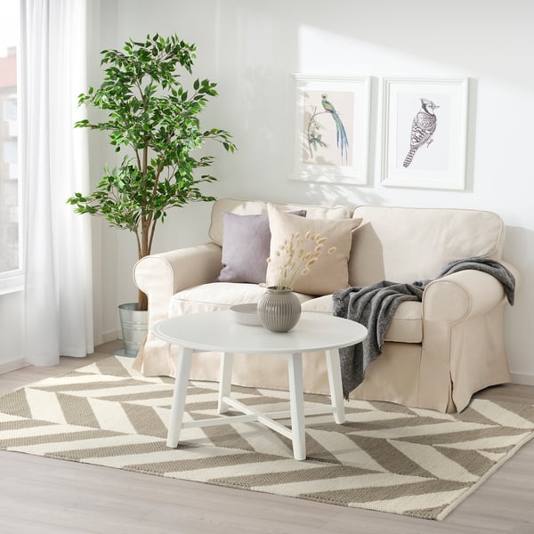 Der LOKSTALL Teppich, beige & grau gestreift, Wolle & Baumwolle, unter einer weißen Couch, neben einem runden weißen Tisch, daneben eine große Pflanze.