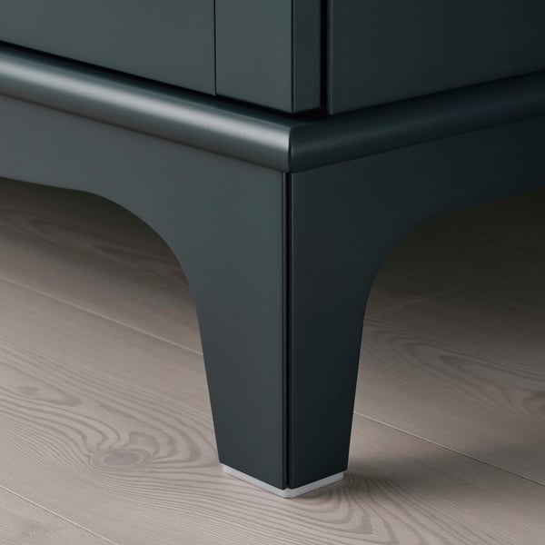 Schlanker, moderner Schrank mit spitz zulaufenden Beinen, dunkelgrüner Oberfläche, hochwertigen Materialien.