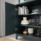 Dunkler Holzschrank mit Glastüren, in denen gestapelte Teller, Weingläser, Kerzen, Vasen und Bücher ausgestellt sind. Sanft schließende Türen und verstellbare Einlegeböden.
