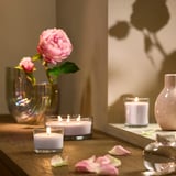 LOTSFÅGEL rosa Duftkerze aus Glas, klein, neben größeren Kerzen und Blumen auf einem Holztisch.