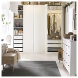 Modernes Wohnzimmer ein weißer Mitteltisch mit Büchern und Vasen, ein Korbstuhl mit Decken, ein gestreifter Teppich und offene Kleiderschränke mit Kleidung.