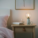 Nachttisch mit hellblauer Lampe, klarer Glühbirne und warmem Licht, teilweise beleuchtetem Buch, Weinglas, in der Nähe des Kopfteils.