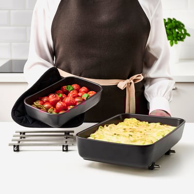 Eine Person in der Schürze hält eine Auflaufform mit gerösteten Tomaten. Davor sitzt ein Auflauf in einer anderen Schale auf einem Untersetzer, beides Teil des LYCKAD-Sets von IKEA.