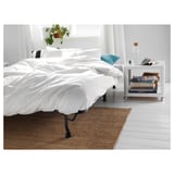 Schlafzimmer mit weißem Bett, schwarzen Beinen, weißem Teppich, Beistelltisch mit Büchern, Flasche und Pflanze.