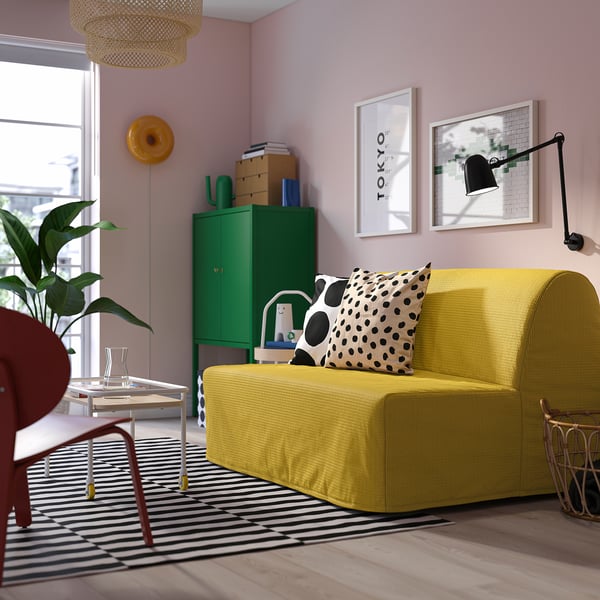 Modernes Wohnzimmer mit gelbem Schlafsofa, schwarz/weißem Teppich, grünem Schrank, farbenfrohem Dekor und zwei Wandbehängen.