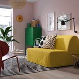 Modernes Wohnzimmer mit gelbem Schlafsofa, schwarz/weißem Teppich, grünem Schrank, farbenfrohem Dekor und zwei Wandbehängen.