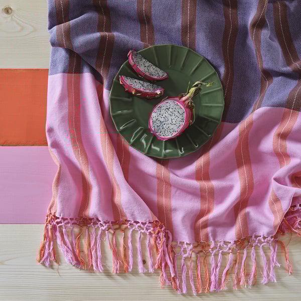 Eine geflochtene Decke mit rosa und blauen Streifen und Fransen steht auf einem Holztisch. Er zeigt zwei Drachenfruchthälften auf einem grünen Teller.