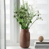 Die handgefertigte und breite Terrakotta-Vase MÄVINN steht neben einem Fenster mit hohen grünen Pflanzen, die auf einem Bücherstapel mit einer Holzkugel ruhen.