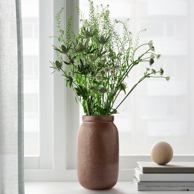 Die handgefertigte und breite Terrakotta-Vase MÄVINN steht neben einem Fenster mit hohen grünen Pflanzen, die auf einem Bücherstapel mit einer Holzkugel ruhen.