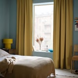 Schlafzimmer mit MAJGULL beige-gelben Vorhängen, großem Fenster, Verdunkelungseffekt. Beigefarbenes Bett, Nachttische aus Holz, blaue Wand.
