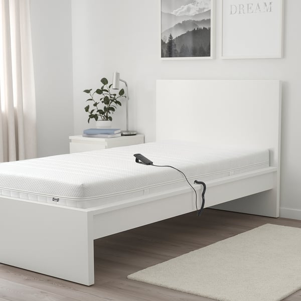 Weiße MAJSKÄR-Schaumstoffmatratze auf dem Bett mit Fernbedienung. Größer als Nachttisch und Teppich, passt auf den Bettrahmen. Pflanze und Lampe auf dem Nachttisch.