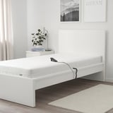 Weiße MAJSKÄR-Schaumstoffmatratze auf dem Bett mit Fernbedienung. Größer als Nachttisch und Teppich, passt auf den Bettrahmen. Pflanze und Lampe auf dem Nachttisch.
