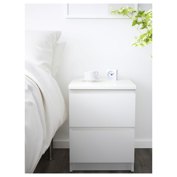 Moderner minimalistischer weißer Nachttisch MALM. Design mit zwei Schubladen, passt unter ein wandmontiertes Regal. In der Nähe des Bettes mit weißen Kissen und Tasse.
