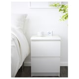 Moderner minimalistischer weißer Nachttisch MALM. Design mit zwei Schubladen, passt unter ein wandmontiertes Regal. In der Nähe des Bettes mit weißen Kissen und Tasse.