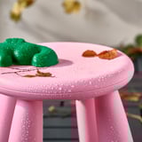 Leuchtend rosa Hocker mit Wassertropfen, grüner Knetmasse und trockenen Blättern obendrauf, perfekt für das Spielen von Kindern im Freien.
