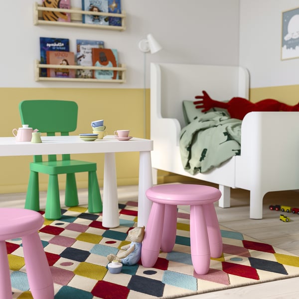 Kinderspielbereich mit grünen MAMMUT-Stühlen, rosa Hockern und weißem Tisch. Heller Teppich, Spielzeug und ungemachtes Bett.
