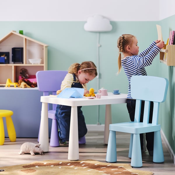 Kinder spielen mit Spielzeug-Teeservice an einem bunten Tisch, einer auf einem blauen Stuhl, der andere auf einem lila Hocker, ein weißes Puppenhaus in der Nähe.