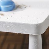 Wassertropfen perlen auf weißer Kindertischkante für den Außenbereich. Der blaue Gegenstand sitzt auf dem Tisch. Perfekt für Picknicks.