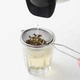 Ein MEDARBETARE Metallsieb sitzt auf einem Glasbecher und filtert Tee. Der Henkel ruht auf dem Becherrand. Edelstahl, passend für verschiedene Tassengrößen.