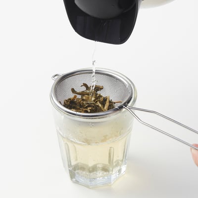 Ein MEDARBETARE Metallsieb sitzt auf einem Glasbecher und filtert Tee. Der Henkel ruht auf dem Becherrand. Edelstahl, passend für verschiedene Tassengrößen.