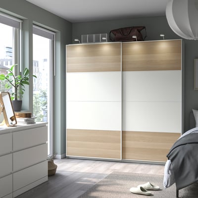 Modernes Schlafzimmer mit hellbraun-weißem MEHAMN Schiebeschrank, Holzboden, grauen Wänden, großem Fenster, gemütlichem Teppich und Bett.