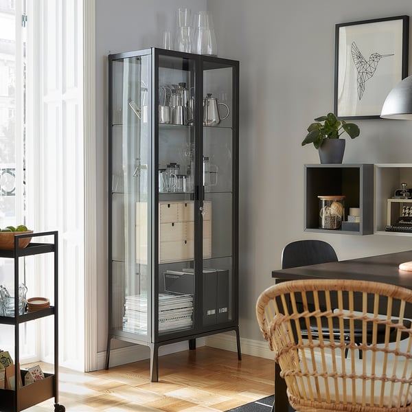 Modernes Wohnzimmer mit schwarzem MILSBO-Glastürschrank mit Glaswaren, Holzstuhl, Topfpflanze und gerahmter Kunst an grauer Wand.