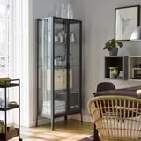 Modernes Wohnzimmer mit schwarzem MILSBO-Glastürschrank mit Glaswaren, Holzstuhl, Topfpflanze und gerahmter Kunst an grauer Wand.