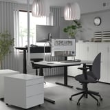 Modernes Büro mit MITTZON Sitz-Steh-Schreibtisch, grauem Stuhl, zwei Bildschirmen, Lampen und Stauraum. Viel Beinfreiheit, verstellbar, kabelgeführt.