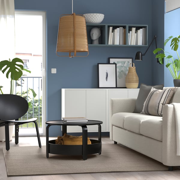 Modernes Wohnzimmer mit blauen Wänden, weißem Sofa, schwarzem Couchtisch, Korblampe, schwarzem Stuhl, Pflanzen, Bücherregal und großem Fenster.