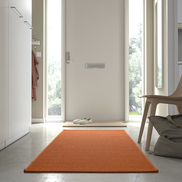 Leuchtend orangefarbener MORUM Teppich von der Tür, passt in den Flur, mit weißen Turnschuhen in der Nähe, modernes, minimalistisches Dekor.