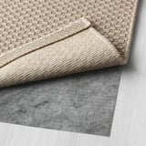 Hellbeiger gewebter Teppich, gefaltet auf dunkelgrauer, rutschfester Unterlage, ausgestellt auf einem hellen Holzboden.