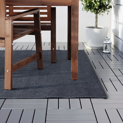 Dunkler Outdoor-Teppich unter hölzernen Terrassenmöbeln. Grauer Belag und weißes Pflanzgefäß sichtbar.
