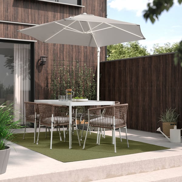 Moderne Terrasse mit grünem MORUM-Teppich, weißem Tisch, grauen Stühlen und großem Sonnenschirm. Holzzaun und Haus im Hintergrund.