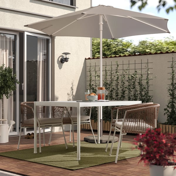 Tisch für den Außenbereich mit weißem Teppich, Korbstühlen, grauem Sonnenschirm, Pflanzen und Haus mit Glastür.