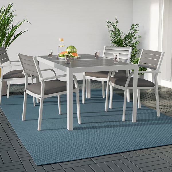 Modernes Esszimmer im Freien mit grauem MORUM-Teppich, weißem Metalltisch und gepolsterten Stühlen. Der Teppich ist groß und erstreckt sich über den größten Teil der Terrasse.