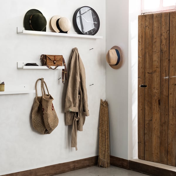 Kleiner Eingangsbereich mit weißen MOSSLANDA Regalen. Hält Hüte, Taschen, einen Mantel und eine schwarze Uhr. Holztür und Stöcke in der Ecke.