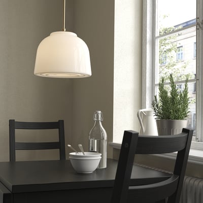 Minimalistische Küchenecke mit schwarzen Möbeln, weißer MUDDERVERK Pendelleuchte, dunklem Tisch, zwei Stühlen, Pflanze und Schale mit Löffeln.