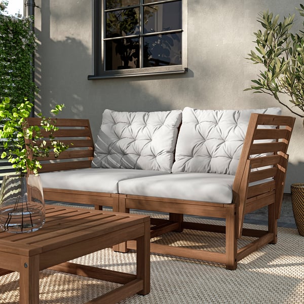 Außenterrasse mit NÄMMARÖ-Möbeln aus brauner Akazie, darunter ein Sofa und ein Couchtisch mit weißen Kissen, auf einem gemusterten Teppich.