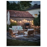 Eine gemütliche Terrasse mit NÄMMARÖ-Holzmöbeln für den Außenbereich mit weißen Kissen, die von warmen Lichterketten und einem geschlossenen Sonnenschirm beleuchtet werden.