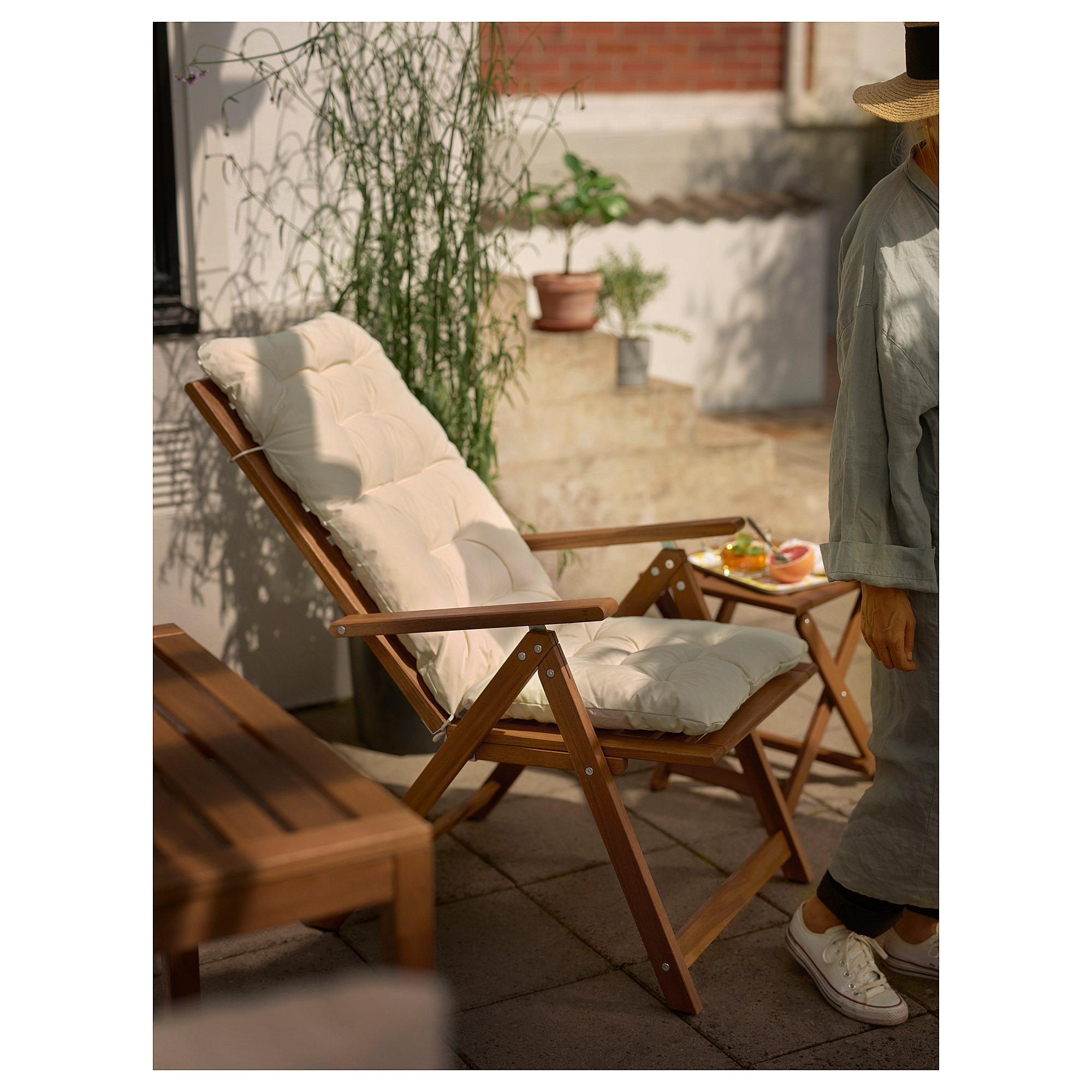 Lounge im Freien mit NÄMMARÖ-Liege, Kissen und kleinem Tisch, umgeben von Topfpflanzen.
